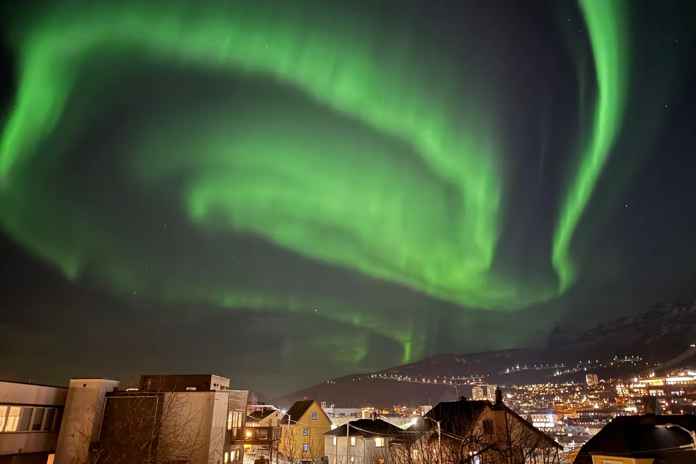 Bright green northern lights arching over the lit houses and hillside lights of Narvik at night