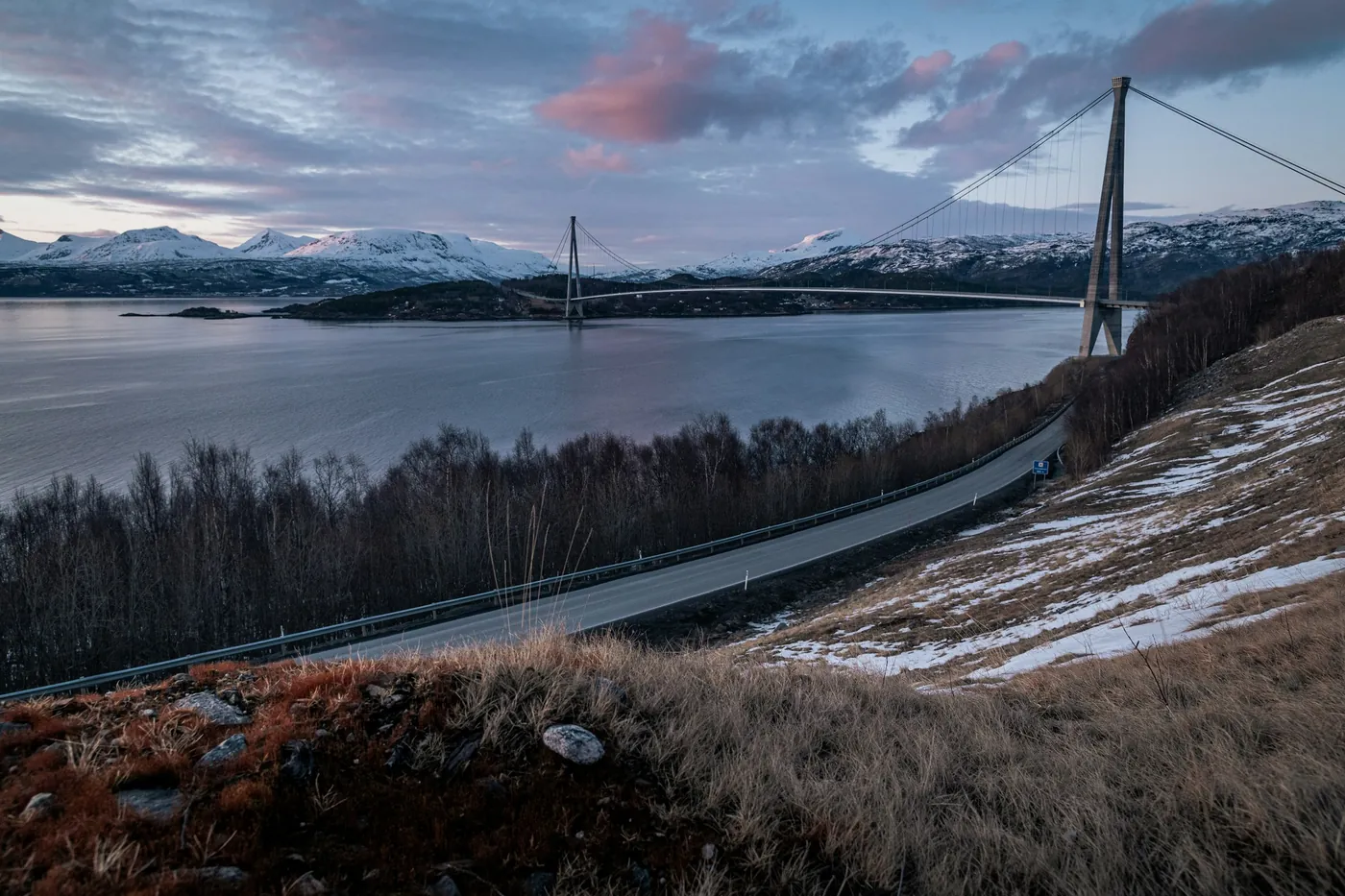 Long suspension bridge spanning a fjord, an approach road curving along a hillside with dry grass and patchy snow, pink clouds