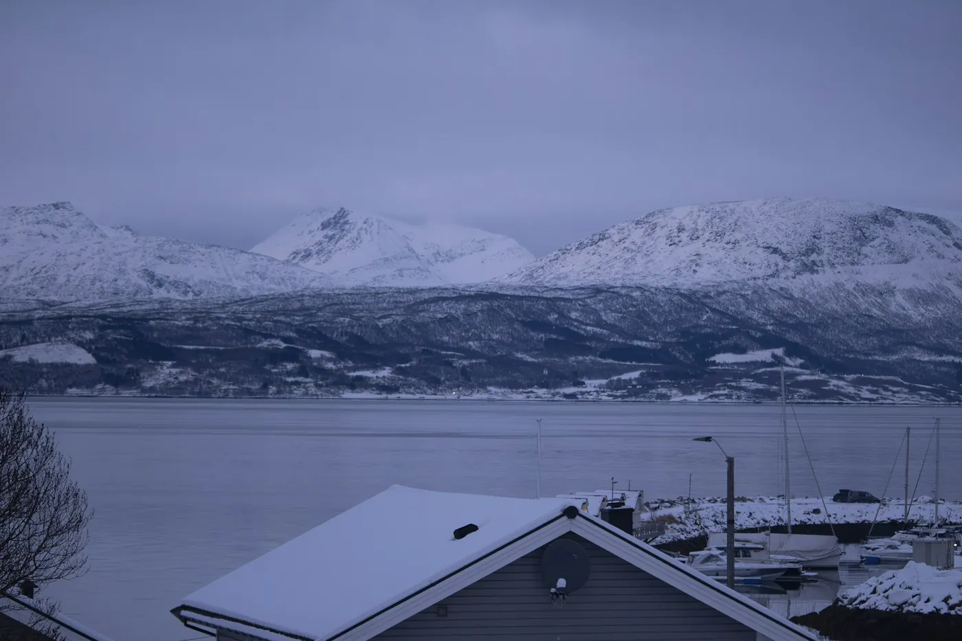 Snow-covered house roofs and a small marina in the foreground, the wide fjord and a chain of snowy mountains behind under a grey-blue winter sky