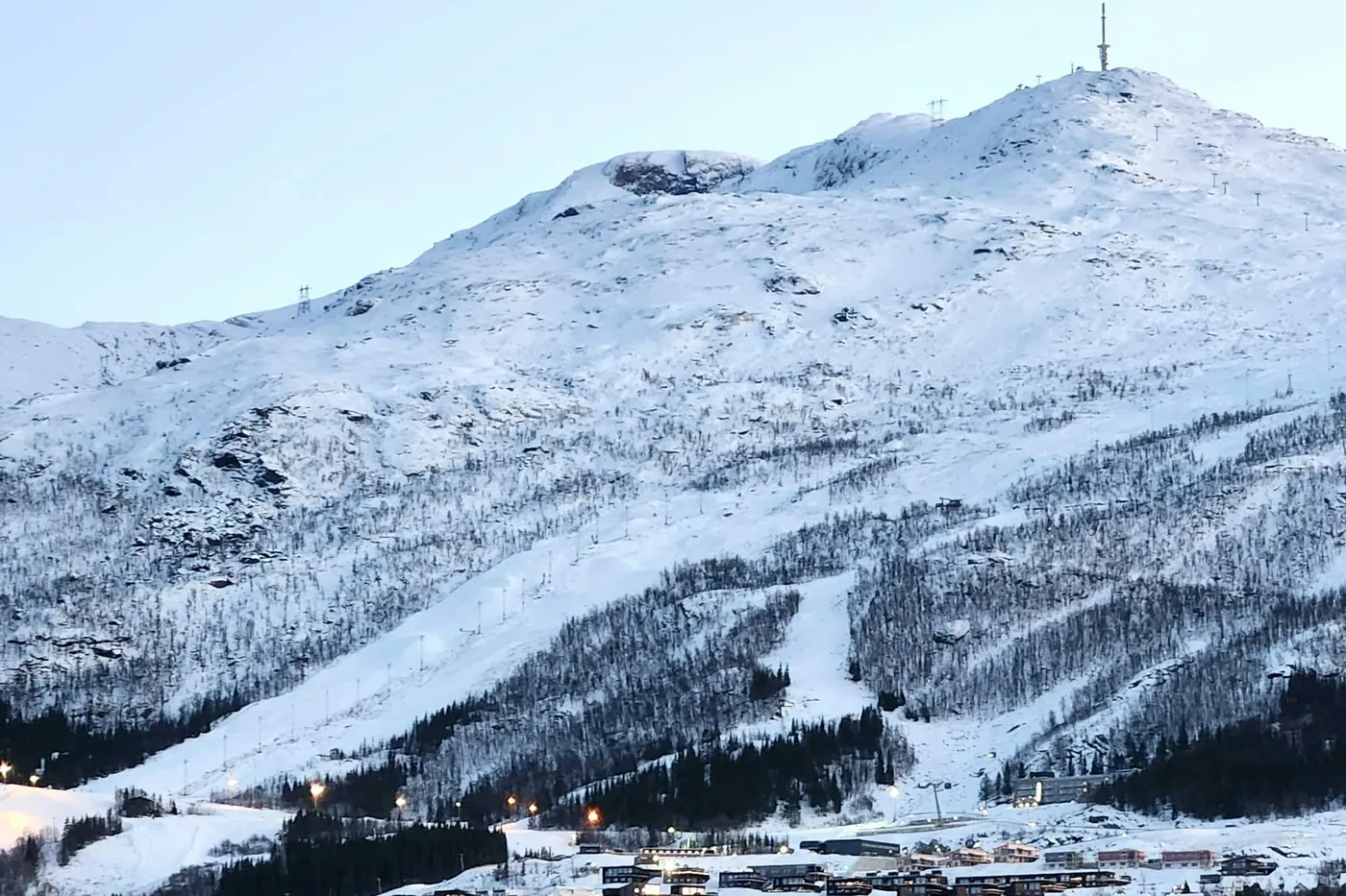 Snowy Narvikfjellet with ski runs, lift pylons and a mast on the summit, town buildings at its foot