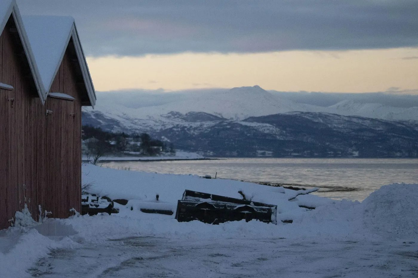 Row of red wooden boathouses with snowy roofs on the shore at Narvik, the fjord and snowy mountains behind under a pale evening sky