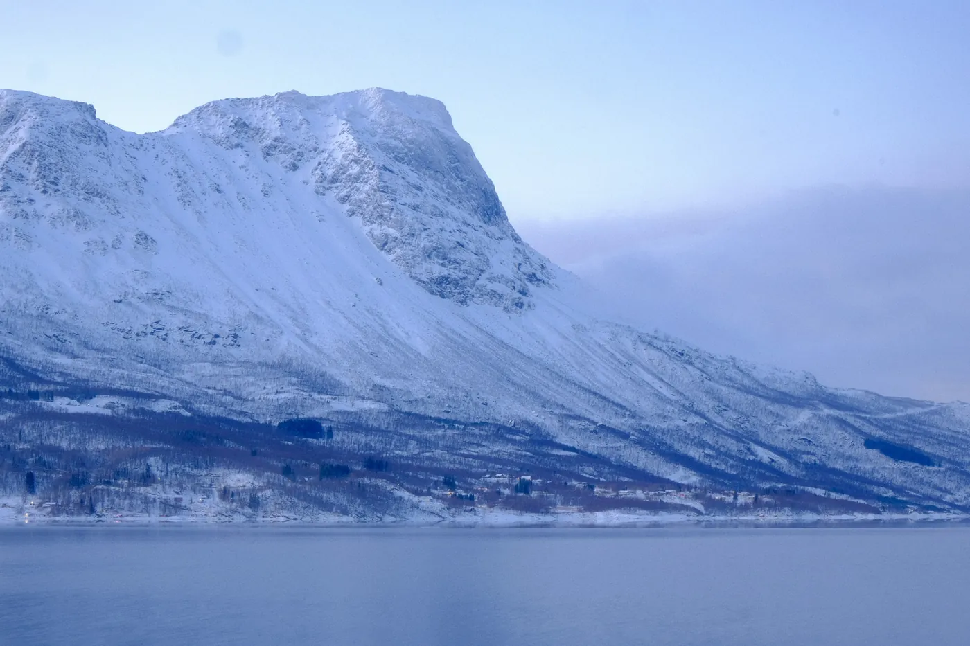 A massive snow-covered mountain rising steeply from calm fjord water near Narvik under a pale blue sky