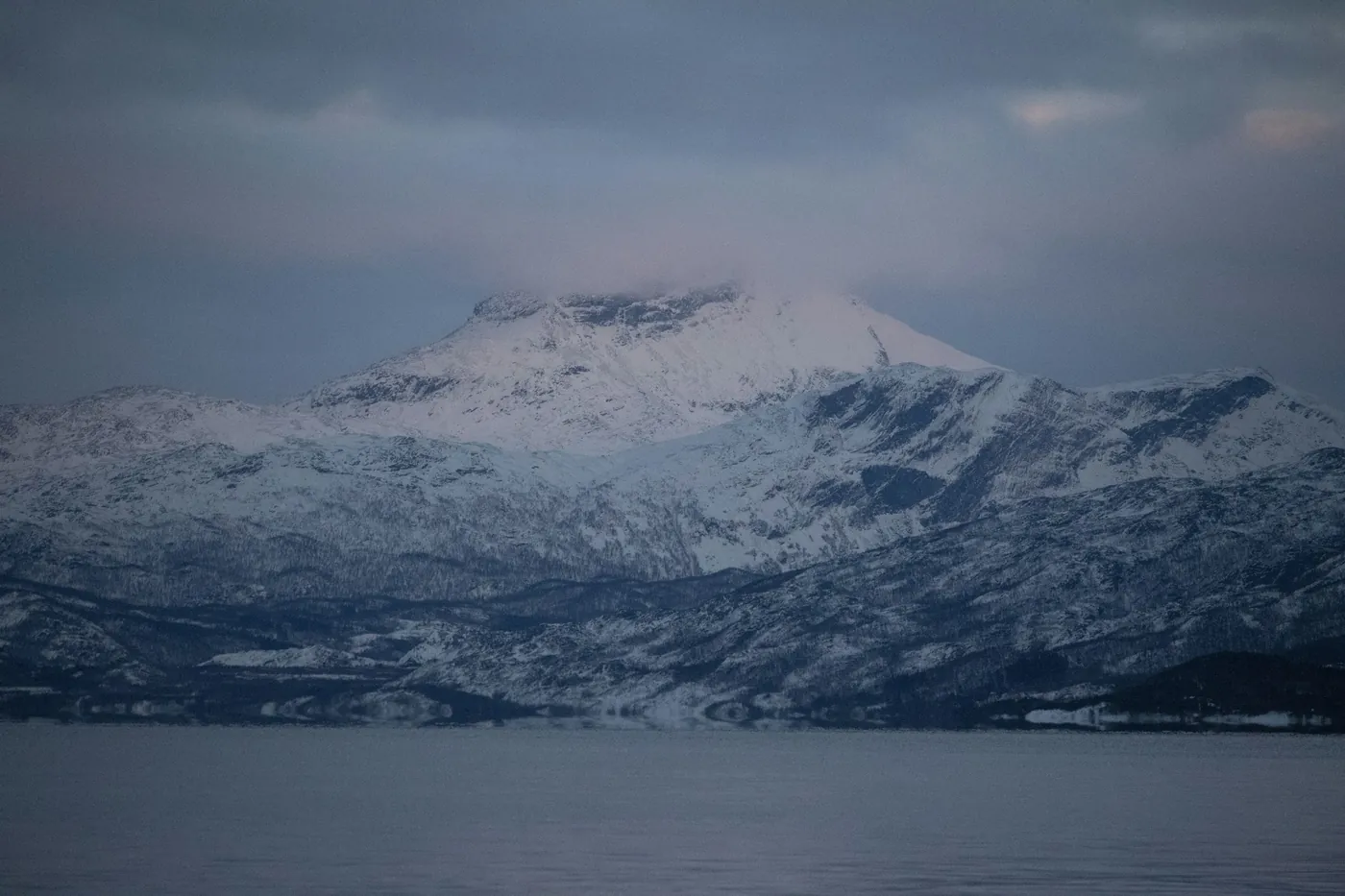 Snow-covered mountains across a dark fjord under heavy grey clouds