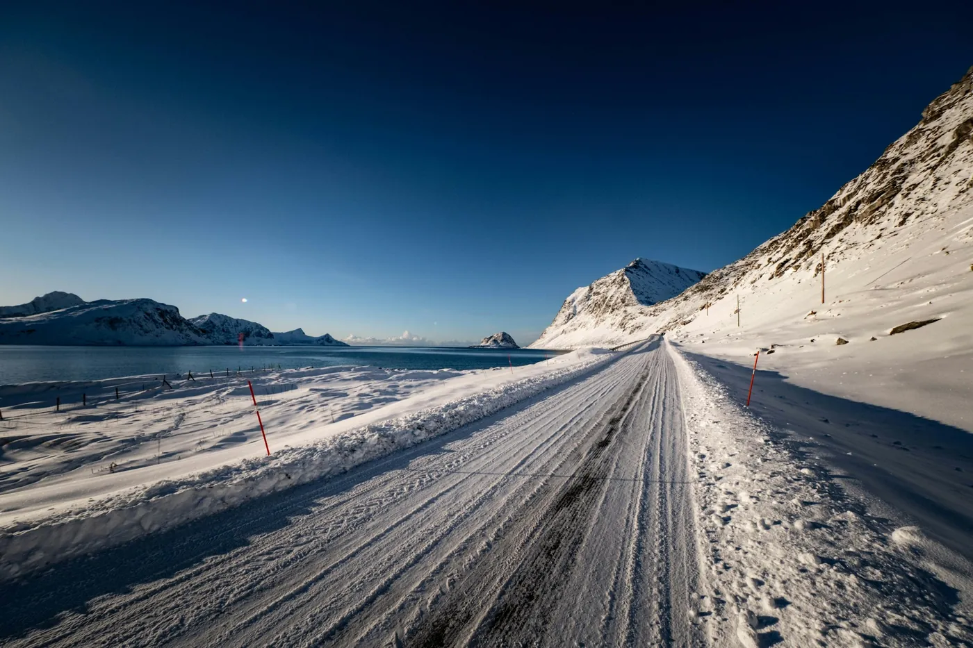 Empty snow-packed coastal road with red marker poles along the sea and snowy hills under a deep blue sky