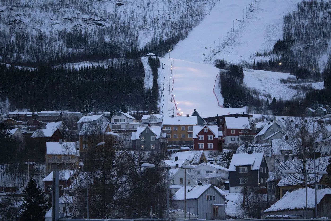 Ski slope lit by floodlights cutting through birch forest above snow-covered houses in Narvik