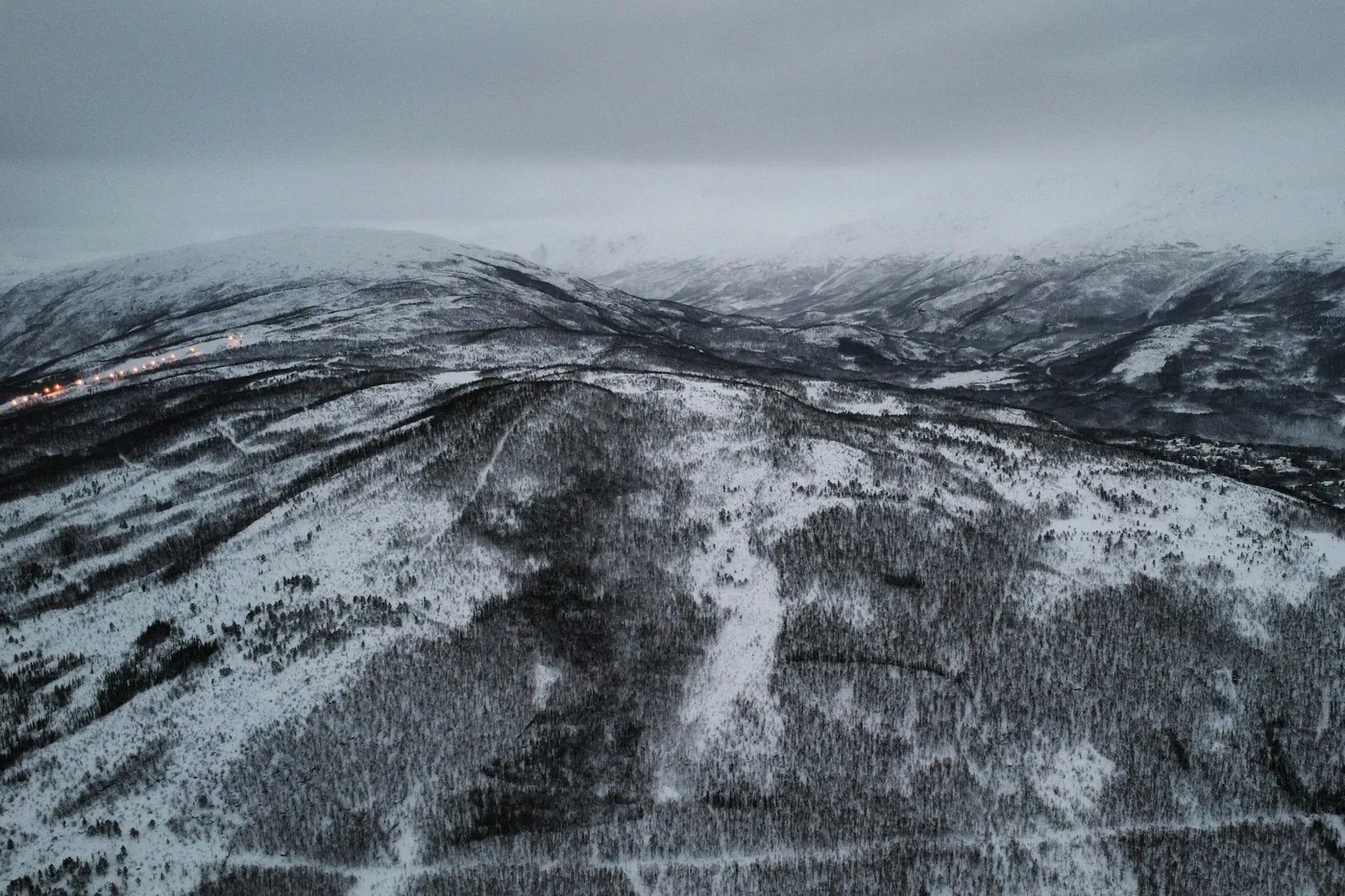 Aerial view over snowy, forested mountain slopes on a grey day, a lit train visible as a line of lights on the far hillside