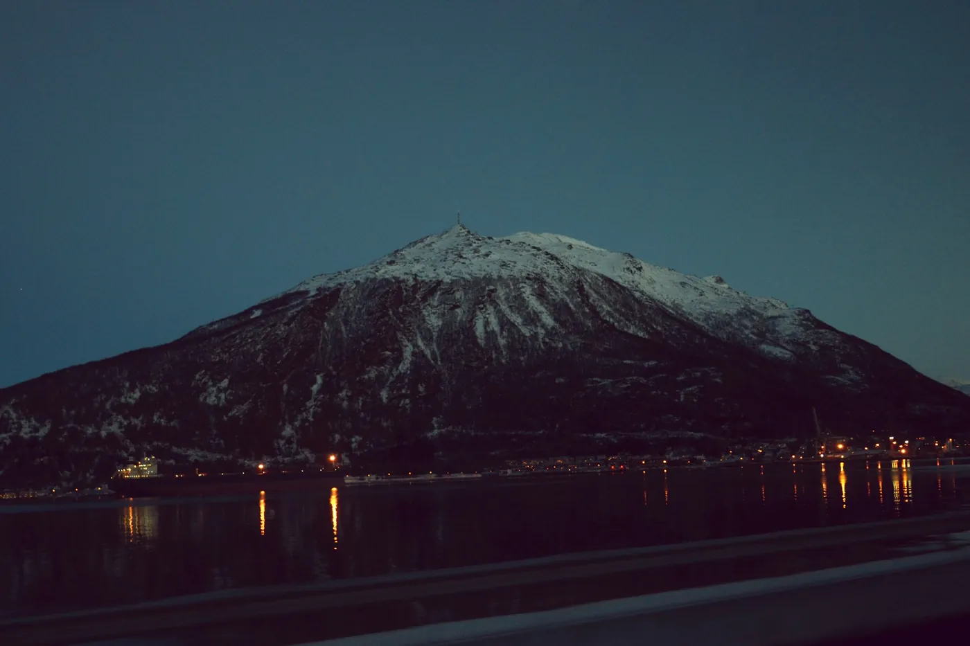 Dark mountain with a snowy summit at dusk above Narvik, town lights reflected in the fjord