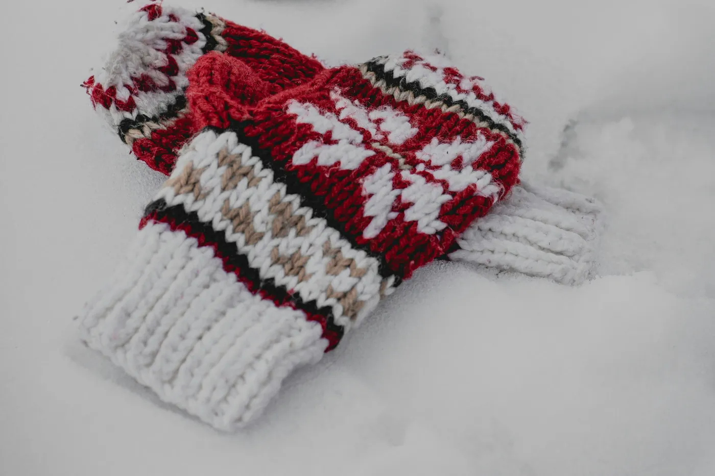 Pair of red and white knitted mittens with a Nordic pattern lying in snow